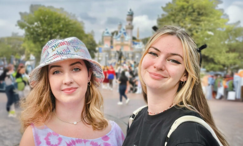 Two young women smiling at the camera in front of Sleeping Beauty Castle at Disneyland. One wears a pastel tie-dye tank top and a bucket hat with “Disneyland” printed across it, while the other wears a black shirt with winged eyeliner and light blonde hair. The castle and park guests are softly blurred in the background.